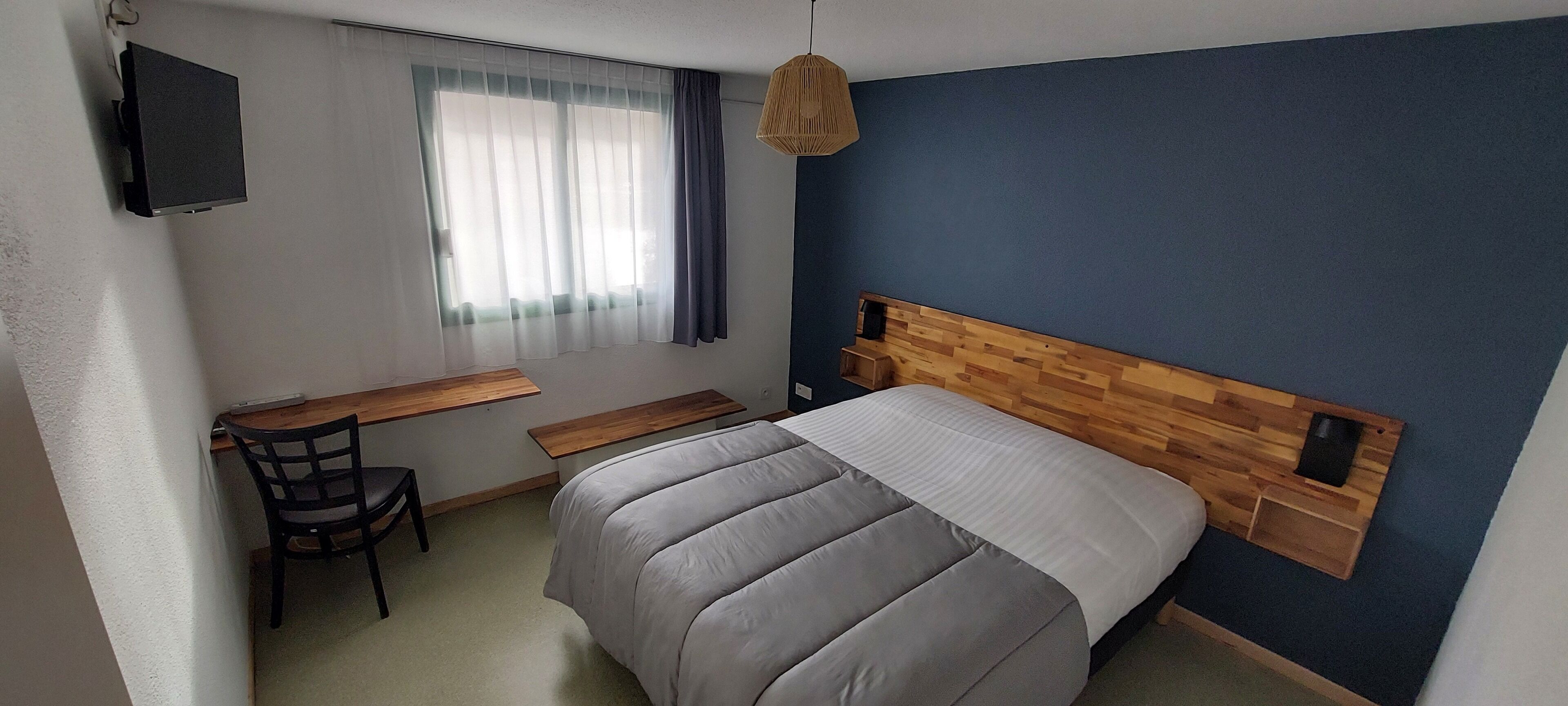 A bedroom with a bed, a desk, a chair, a TV, and a window with curtains.