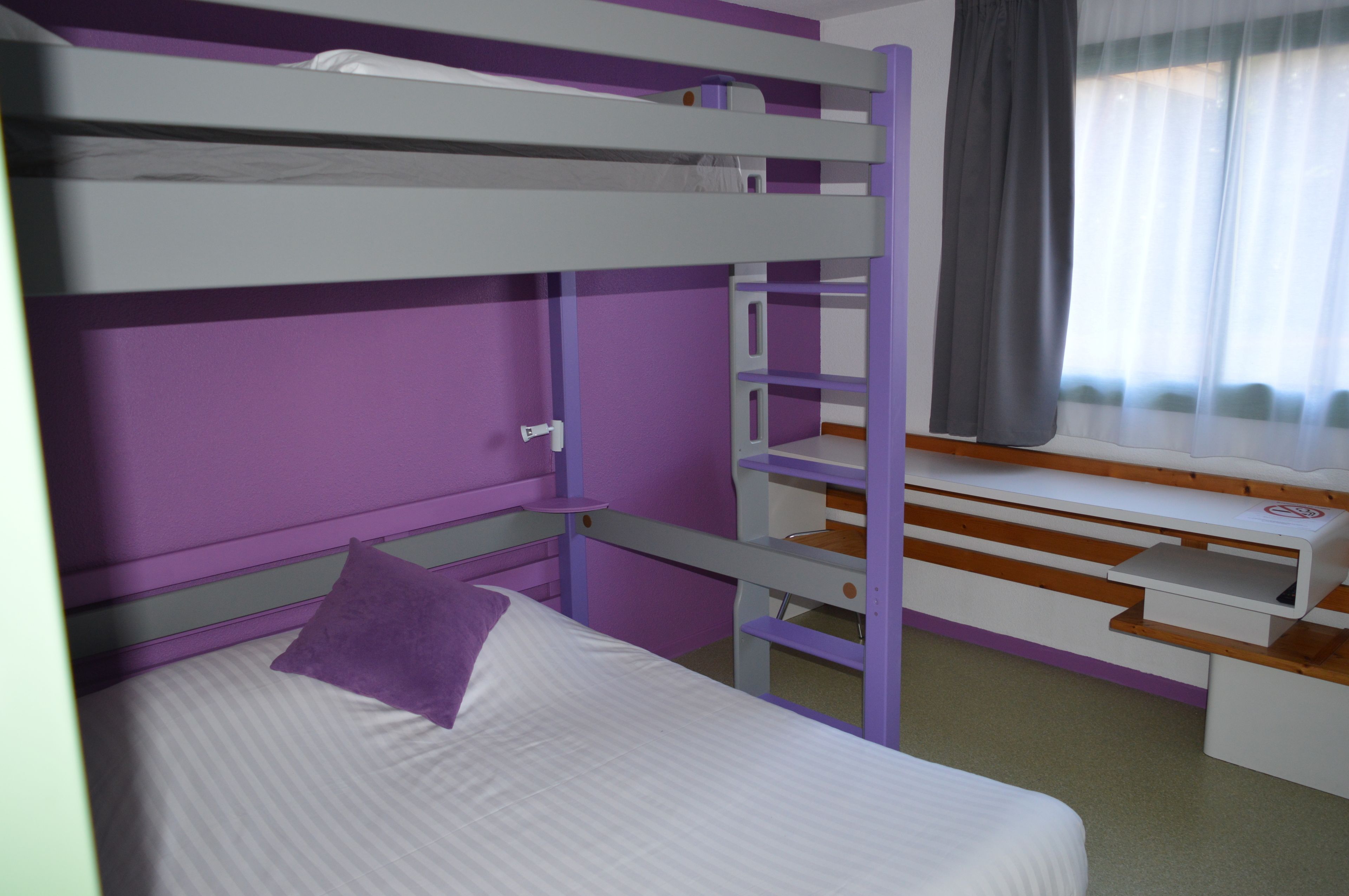 Una cama individual superpuesta con un almohadón morado, un escritorio con una silla y una ventana con cortinas.