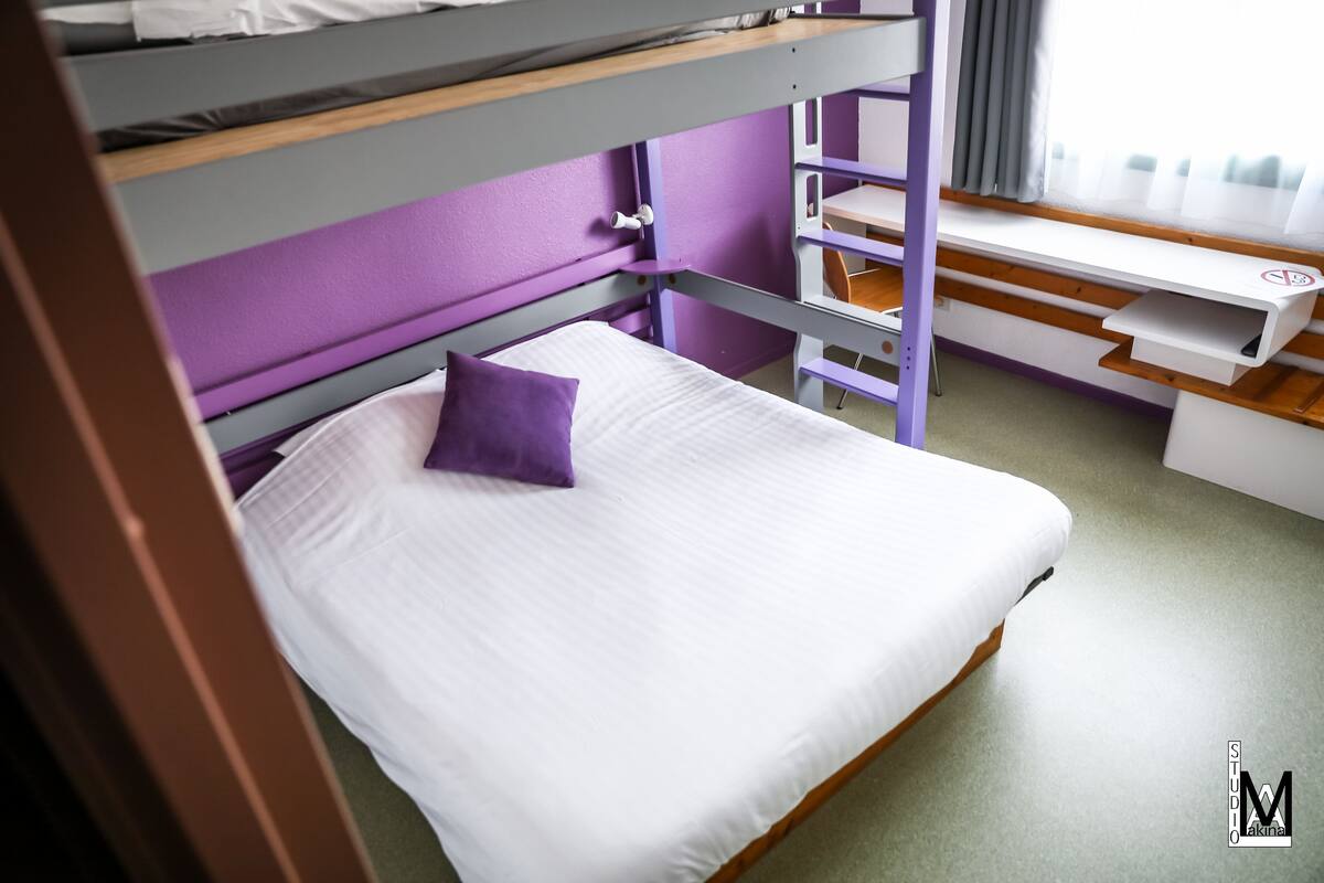 Uma cama beliche com colchão branco e travesseiro roxo, uma escrivaninha com cadeira e uma janela com cortinas.