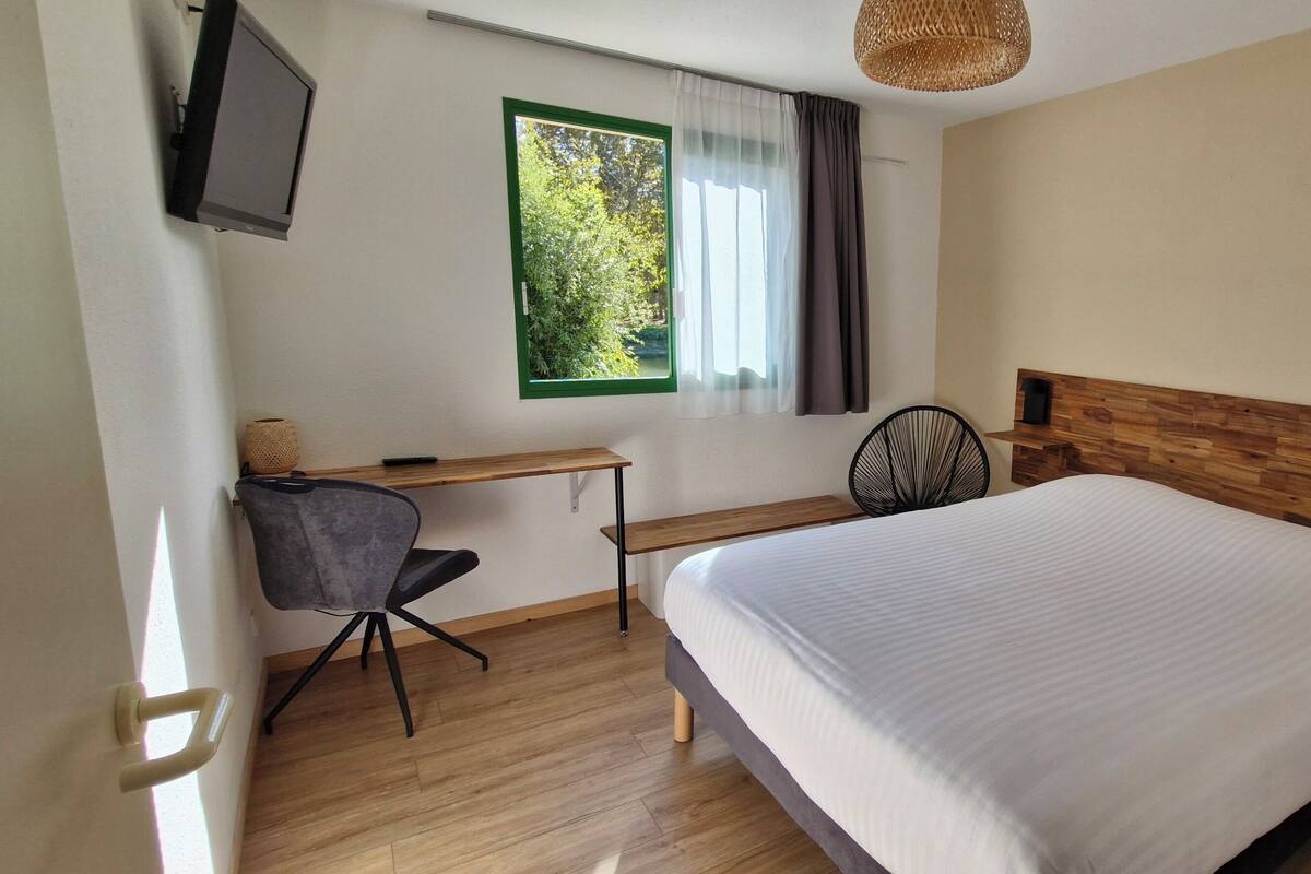 Quarto com cama, escrivaninha, cadeira, televisão e janela com cortinas.