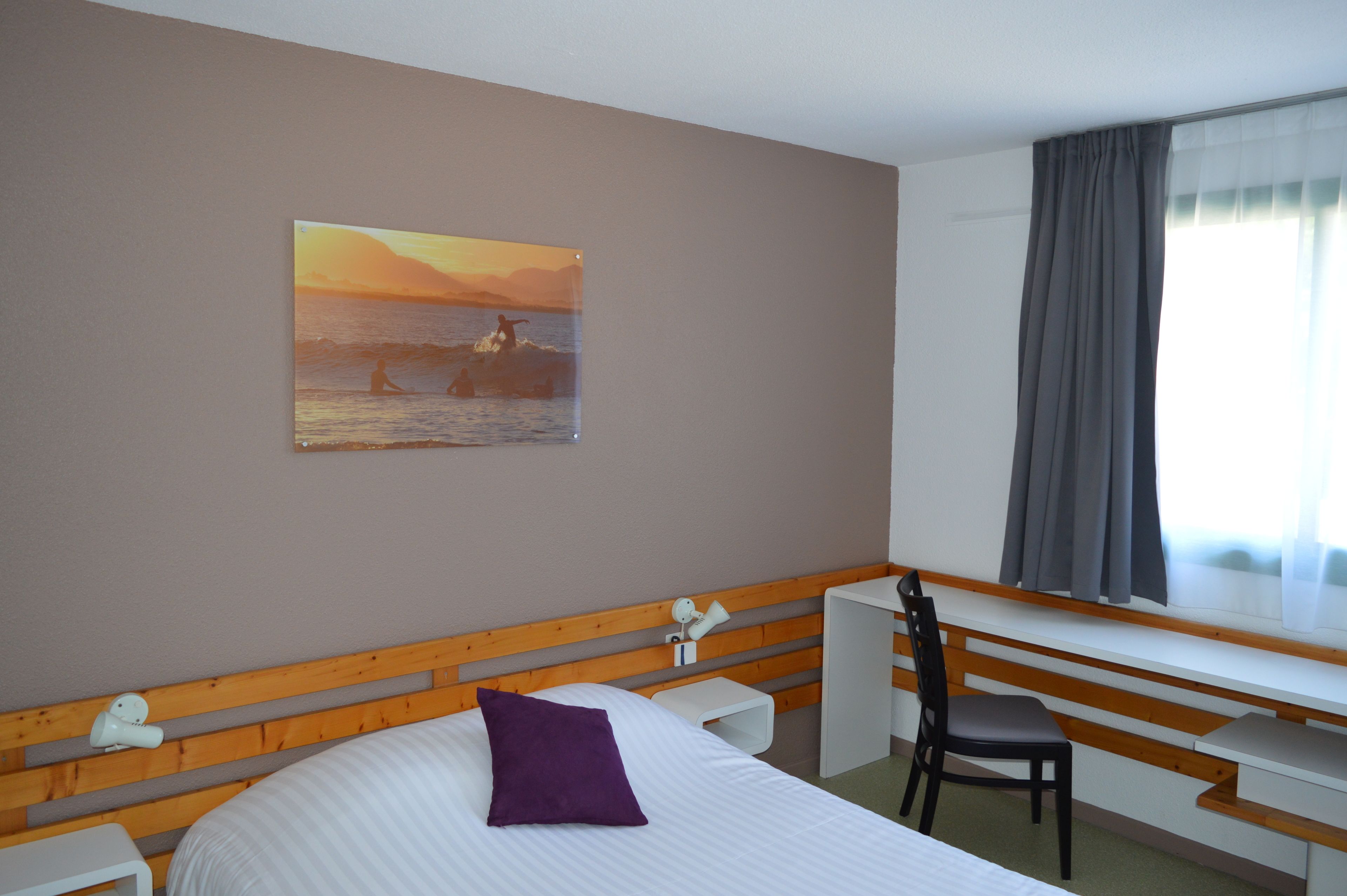 A hotel room with a bed, a desk, a chair, a window with curtains, and a painting on the wall.
