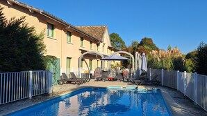 En swimmingpool med solstolar och parasoller, omgiven av ett vitt staket, och en byggnad med ett tak täckt av tegelpannor och gröna fönsterramar.