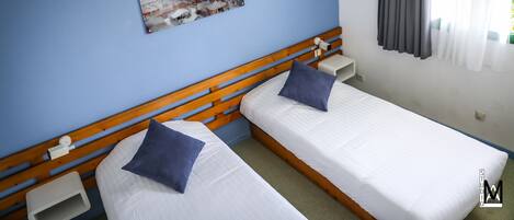 Habitación de hotel con dos camas individuales, cabecera de madera, mesita de noche y televisor montado en la pared, además de una ventana con cortinas.