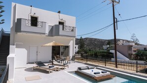 A modern two-story building with a swimming pool, outdoor seating, and a view of the surrounding landscape. - Matala Bay Apartments (Faistos)