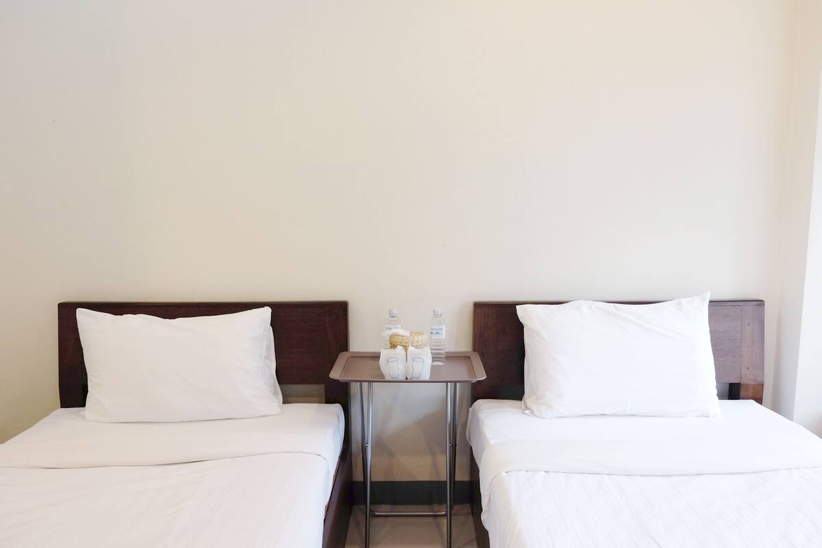 A hotel room with two single beds, a small table with a vase and bottles, and plain walls.