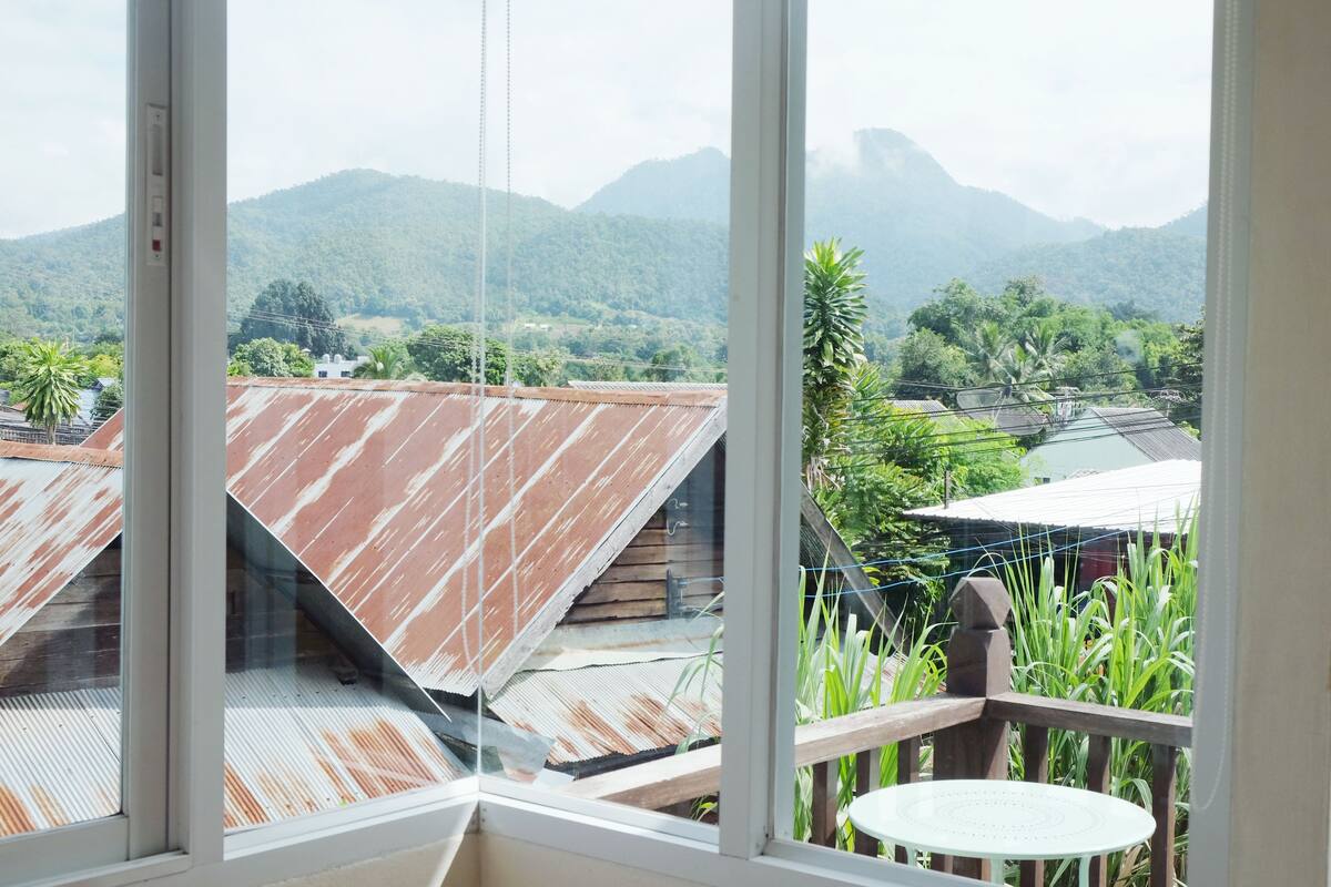 A view from a window with a wooden balcony, overlooking a mountainous landscape with green vegetation and buildings with red roofs.