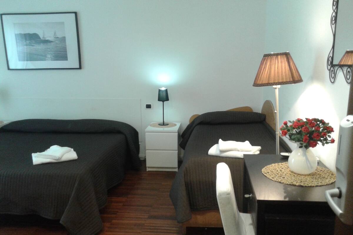 Habitación de hotel con dos camas, una mesita de noche con lámpara, un escritorio con un jarrón de flores y un cuadro en la pared.