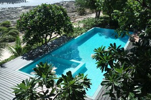 Une piscine rectangulaire dotée d’une terrasse en bois, entourée d’une végétation luxuriante et offrant une vue sur une côte rocheuse.