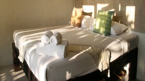 Ein ordentlich gemachtes Bett mit weißen Leinen und aufgerollten Handtüchern, einem grünen Kissen und einem braunen Kissen auf einem hölzernen Bettgestell in einem Raum mit Backsteinwand.