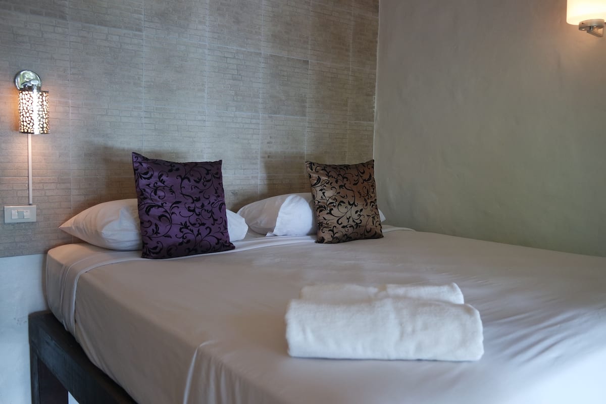 Een net opgemaakt bed met witte lakens en twee gekleurde kussens, een wandlamp en een effen muur.