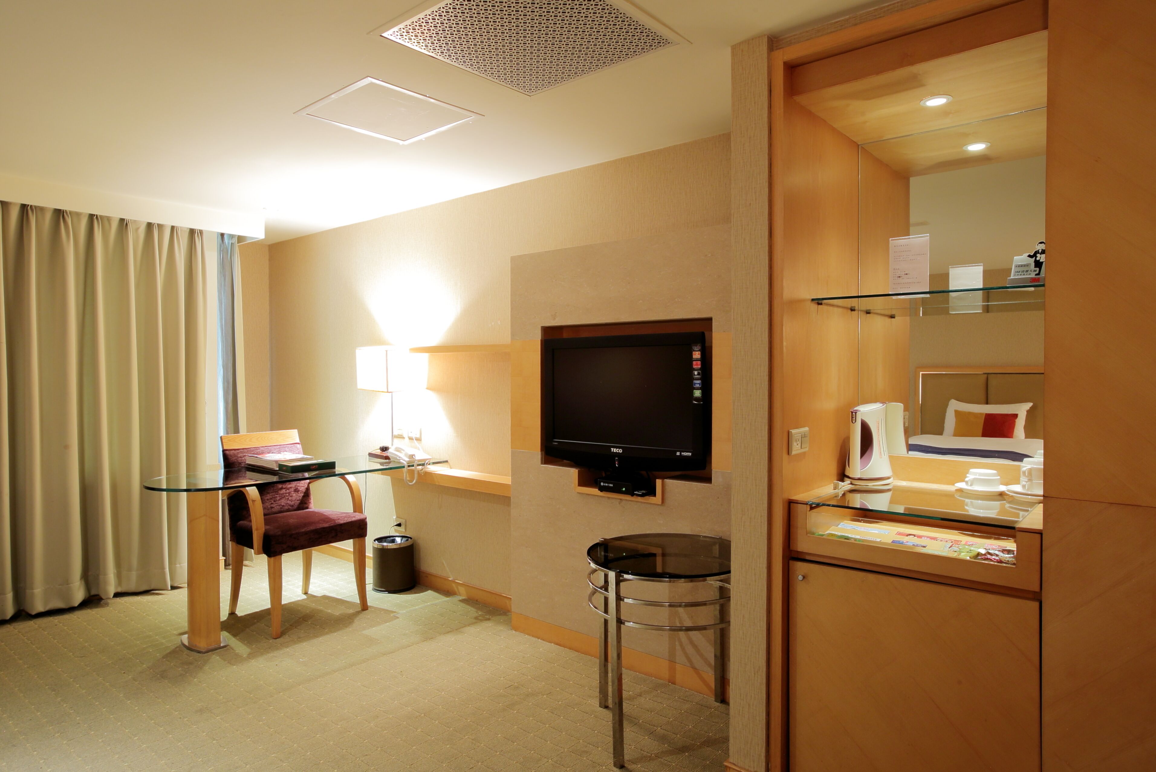 A hotel room with a TV, a glass table, a chair, a lamp, a trash bin, and a minibar.
