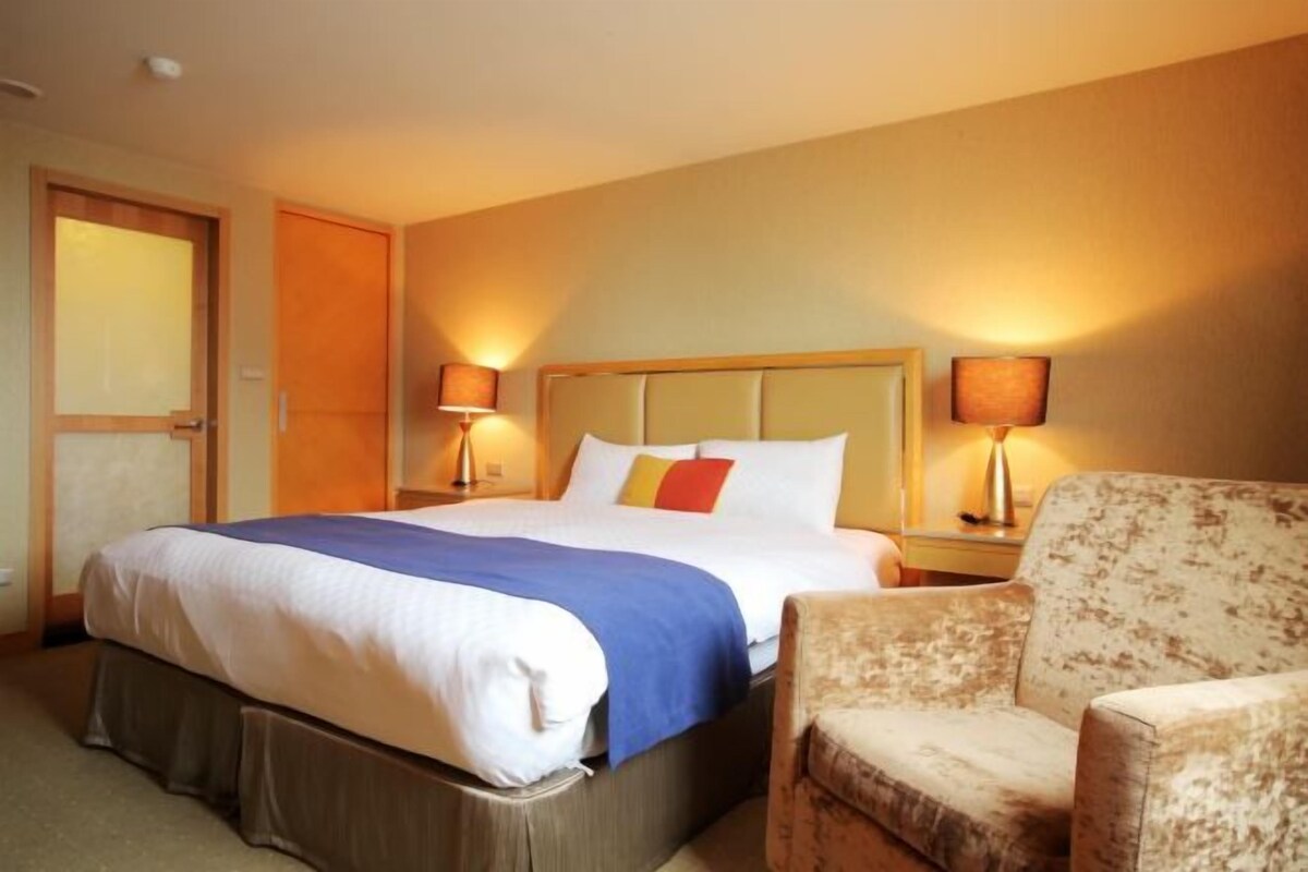 A hotel room with a large bed, two bedside lamps, a chair, and a door.