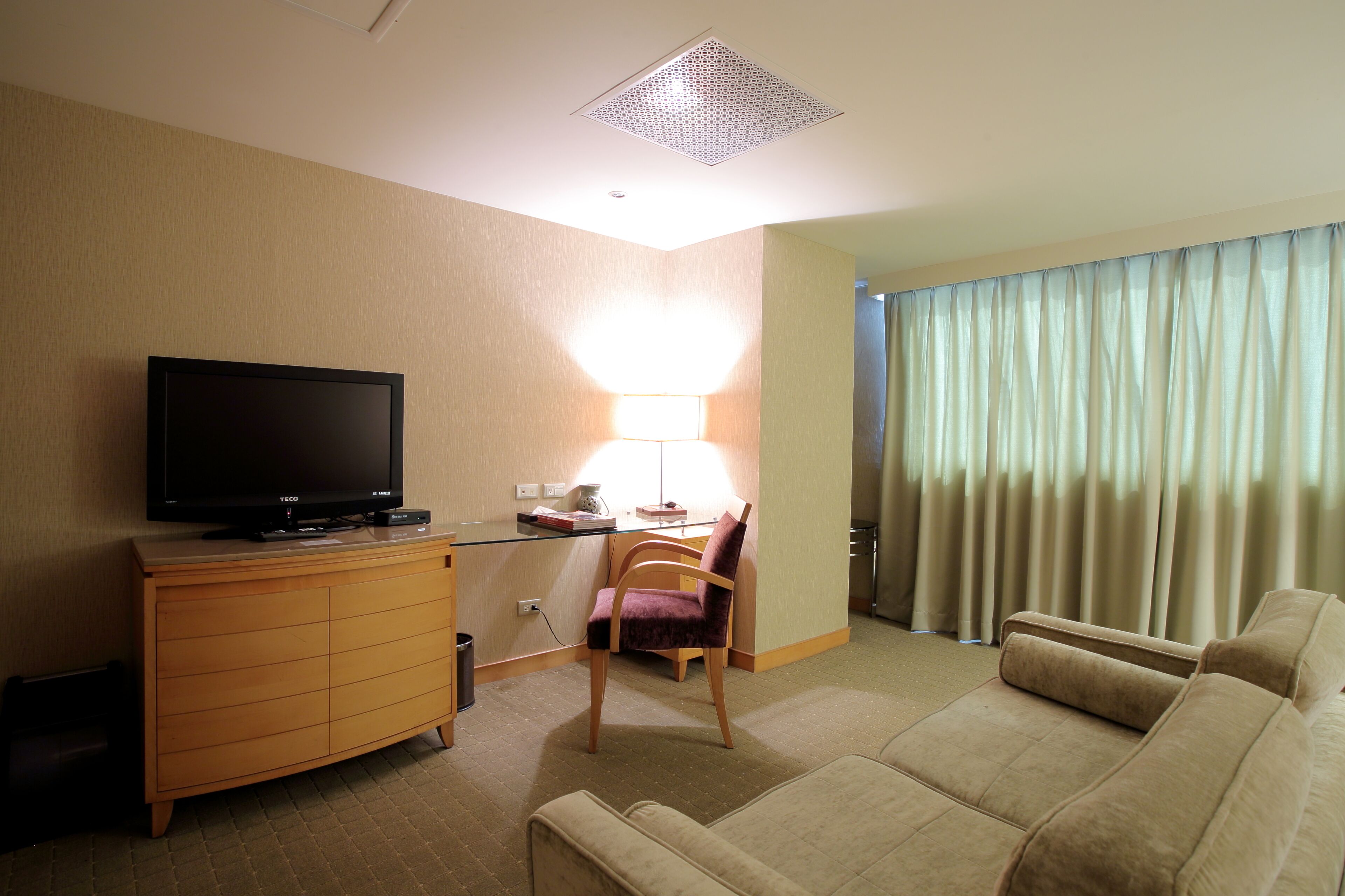 A hotel room with a flat-screen TV on a wooden cabinet, a desk with a lamp, a purple chair, a beige sofa, and a large window with curtains.