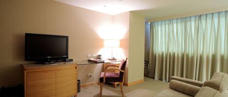 A hotel room with a flat-screen TV on a wooden cabinet, a desk with a lamp, a purple chair, a beige sofa, and a large window with curtains.