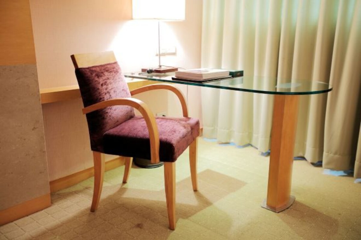 Une chaise avec un siège rembourré et des pieds en bois, une table en verre avec un livre dessus, et une fenêtre avec des rideaux.