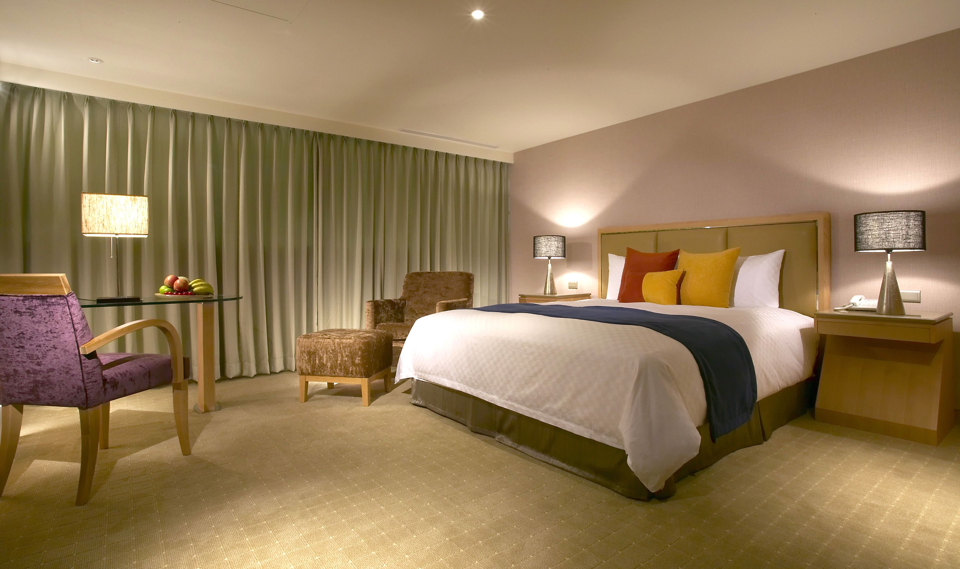 Une chambre d’hôtel avec un grand lit, deux lampes de chevet, une chaise, une petite table avec des fruits et un rideau.