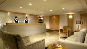 Un espace d’accueil d’hôtel comprenant un comptoir de réception, deux employés, un présentoir, un coin salon avec des fauteuils et un canapé, une petite table avec une bougie et des fleurs, et une horloge murale.