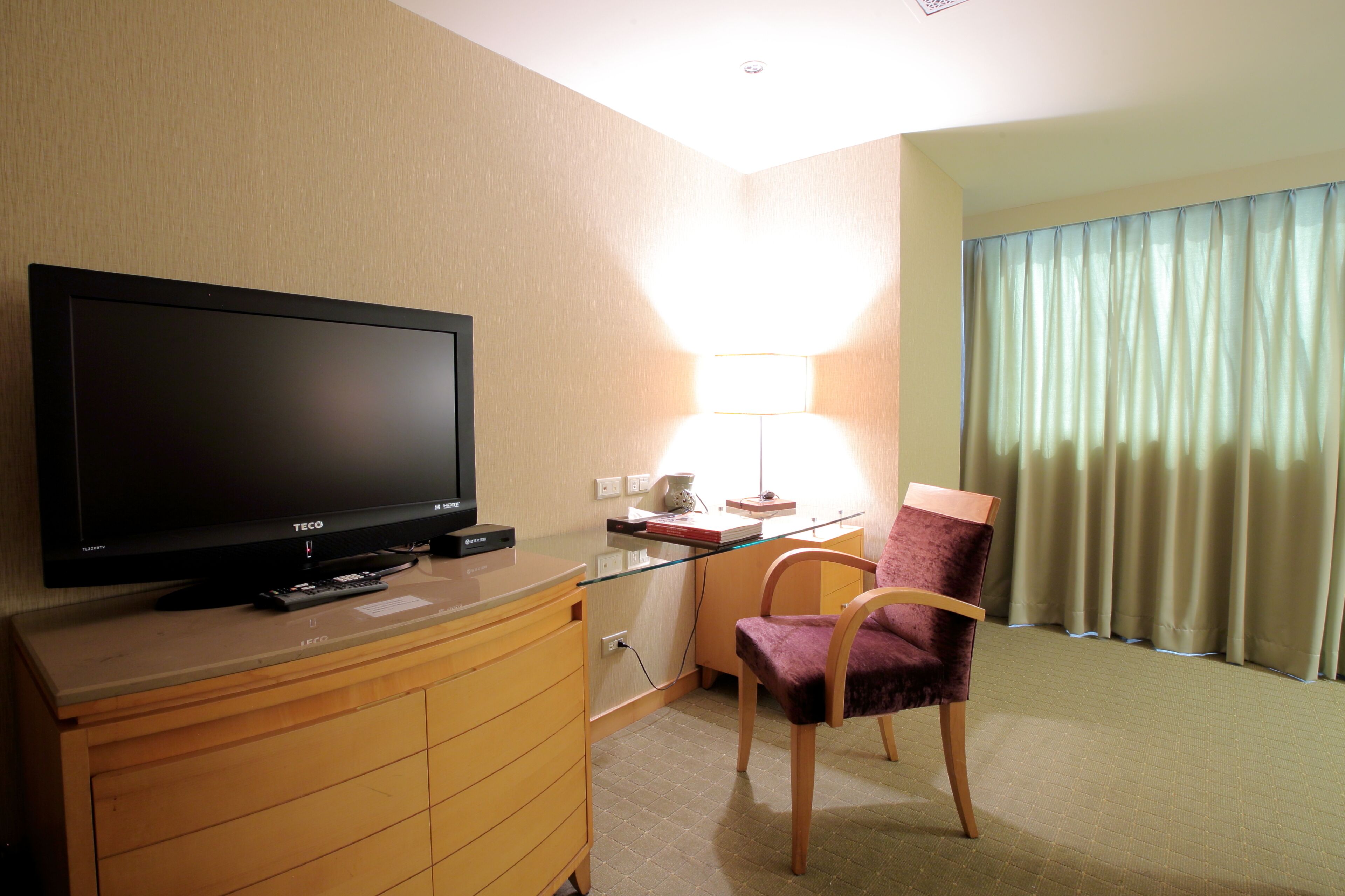 A hotel room with a flat-screen TV on a wooden dresser, a glass desk with a lamp, a purple chair, and a window with curtains.