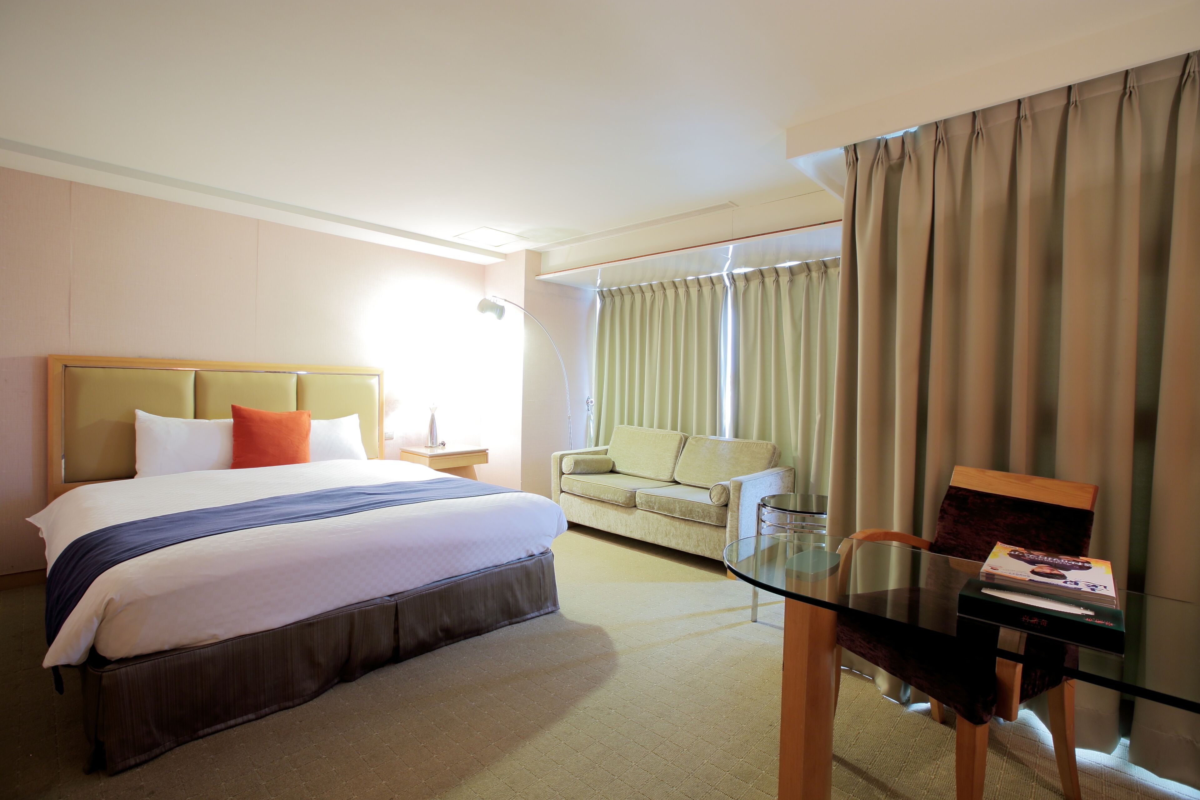A hotel room with a bed, a sofa, a glass table, and a window with curtains.