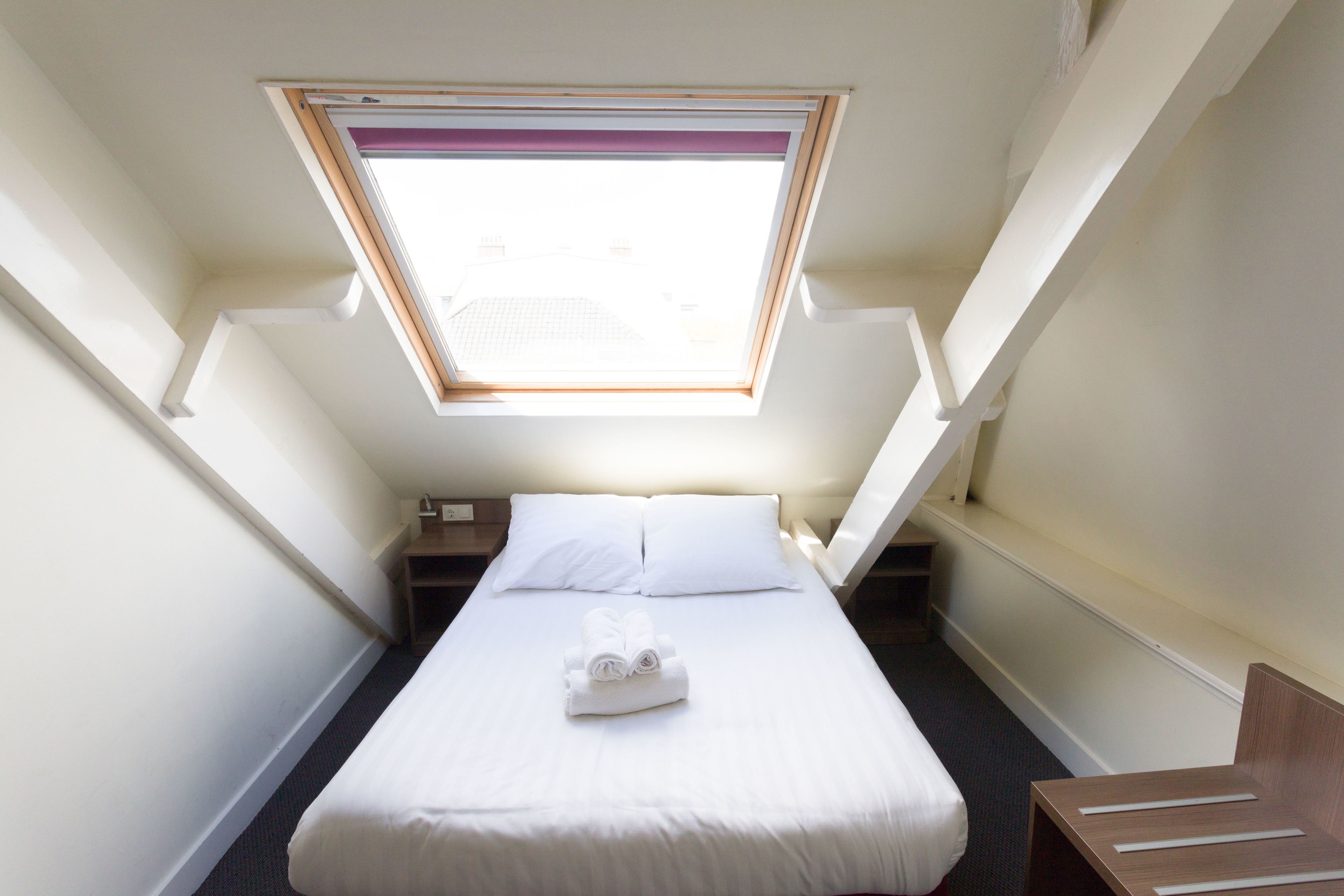 A small, narrow hotel room with a single bed, a nightstand, a wooden chair, and a skylight.