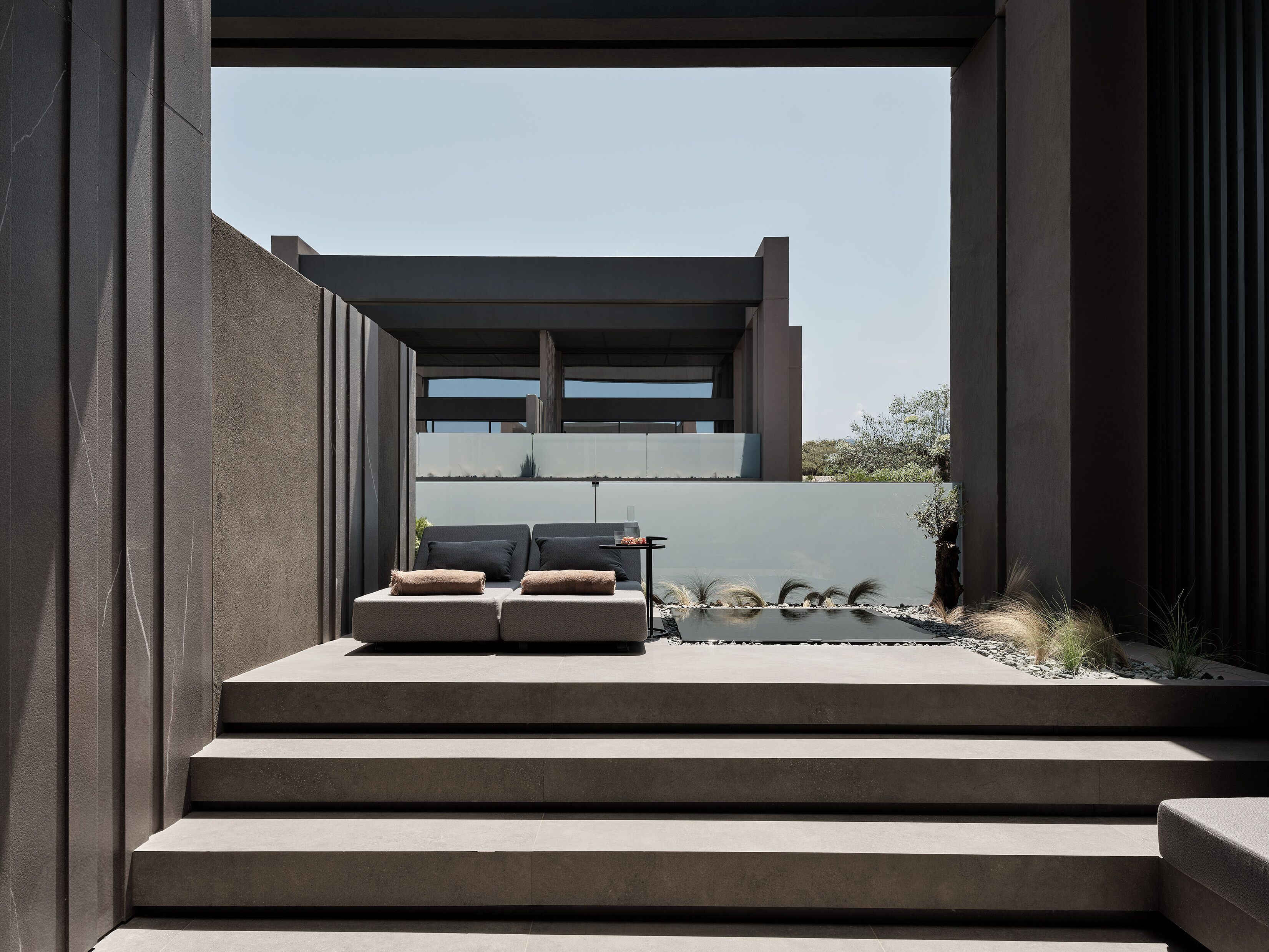 Un área de estar moderna al aire libre con piso de concreto, escalones de acceso y vista de un edificio al fondo.