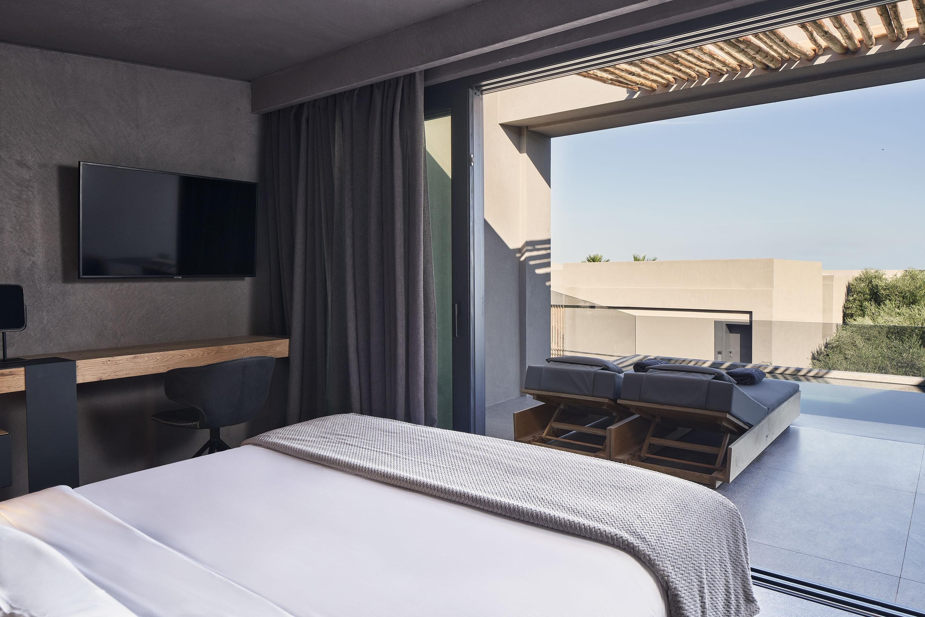 Ein modernes Hotelzimmer mit einem großen Bett, einem Flachbildfernseher an der Wand, einem Schreibtisch mit Stuhl und einem Balkon mit Sitzgelegenheit.