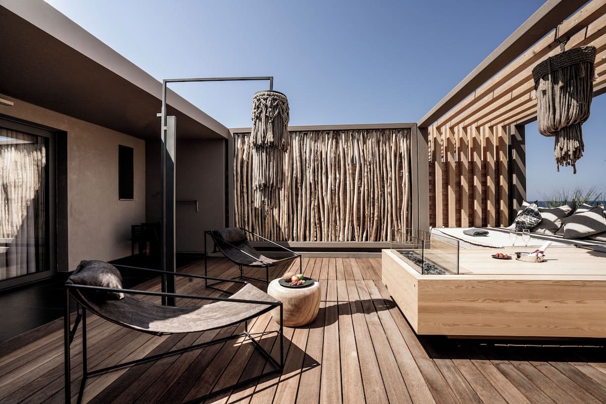 A modern outdoor deck with wooden flooring, a lounge chair, a round ottoman, a hanging basket, and a wooden wall with vertical slats.