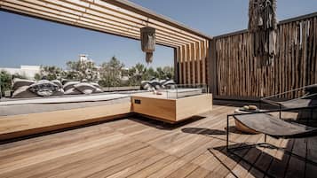 A modern outdoor deck with wooden flooring, a seating area with cushions, a hanging light fixture, and a view of the cityscape.
