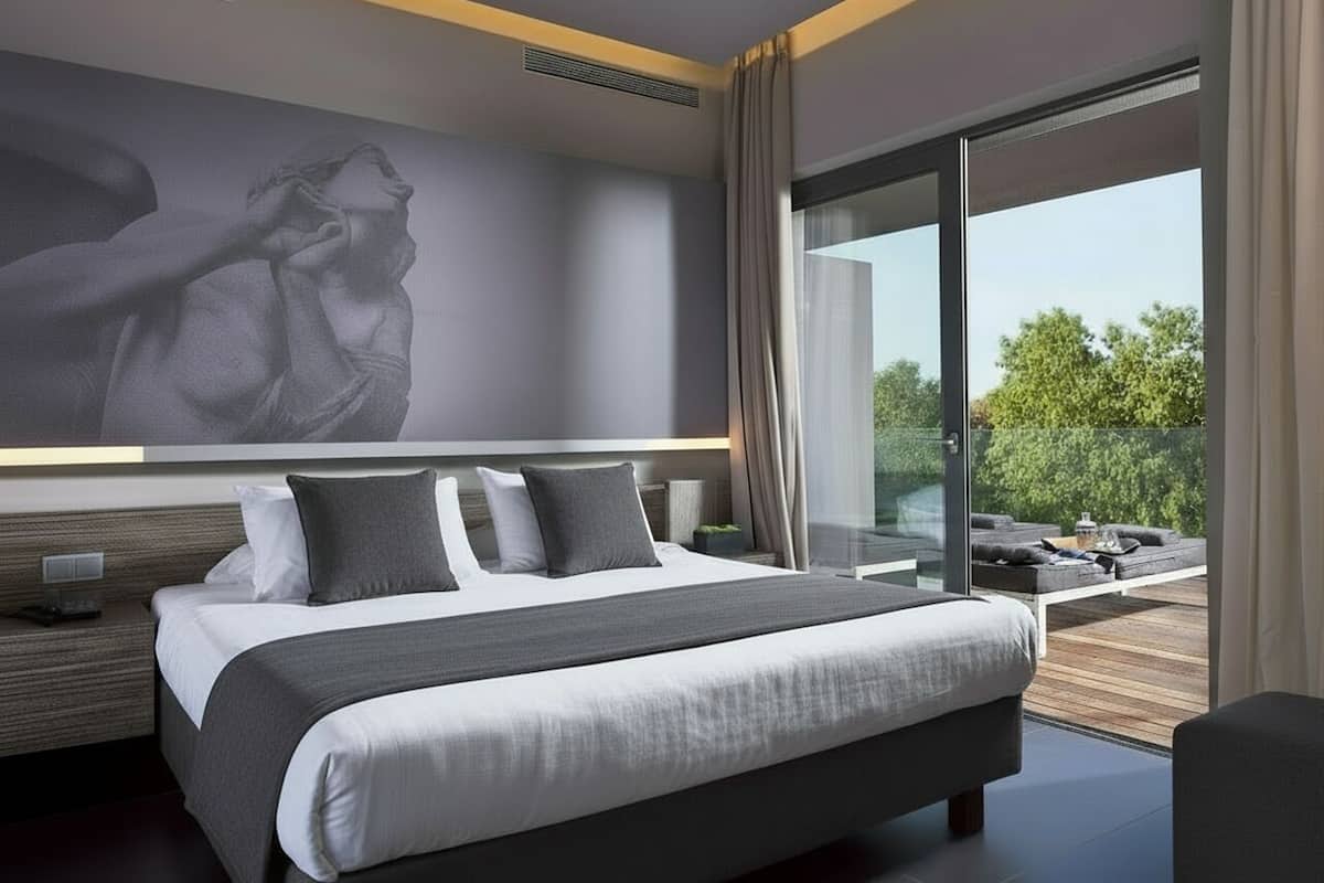 A modern bedroom with a large bed, a nightstand, a wall-mounted light, and a view of the outdoors.