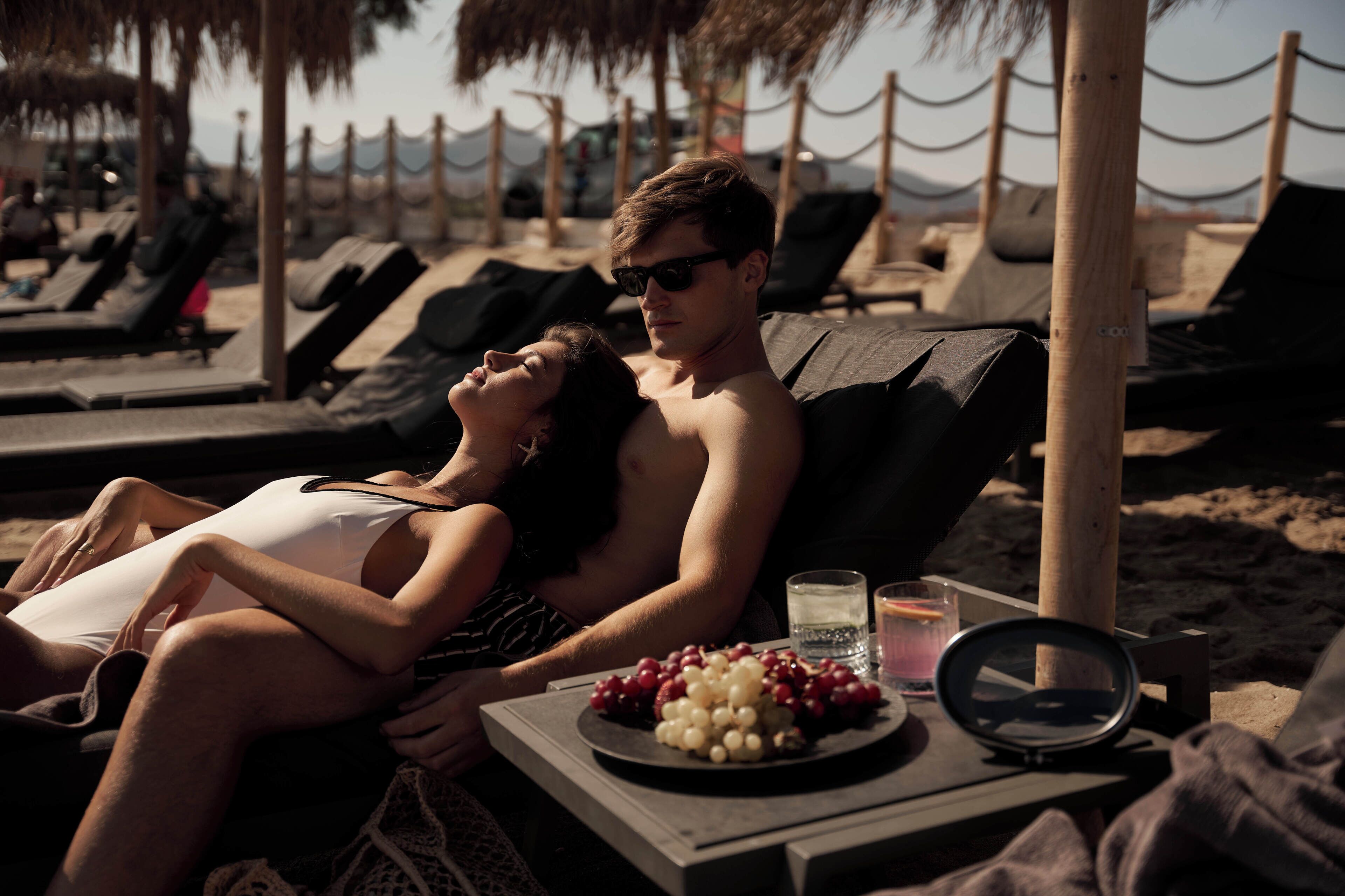 Una pareja descansando en una silla de playa con una mesa frente a ellos que tiene un plato de uvas y un vaso de agua.