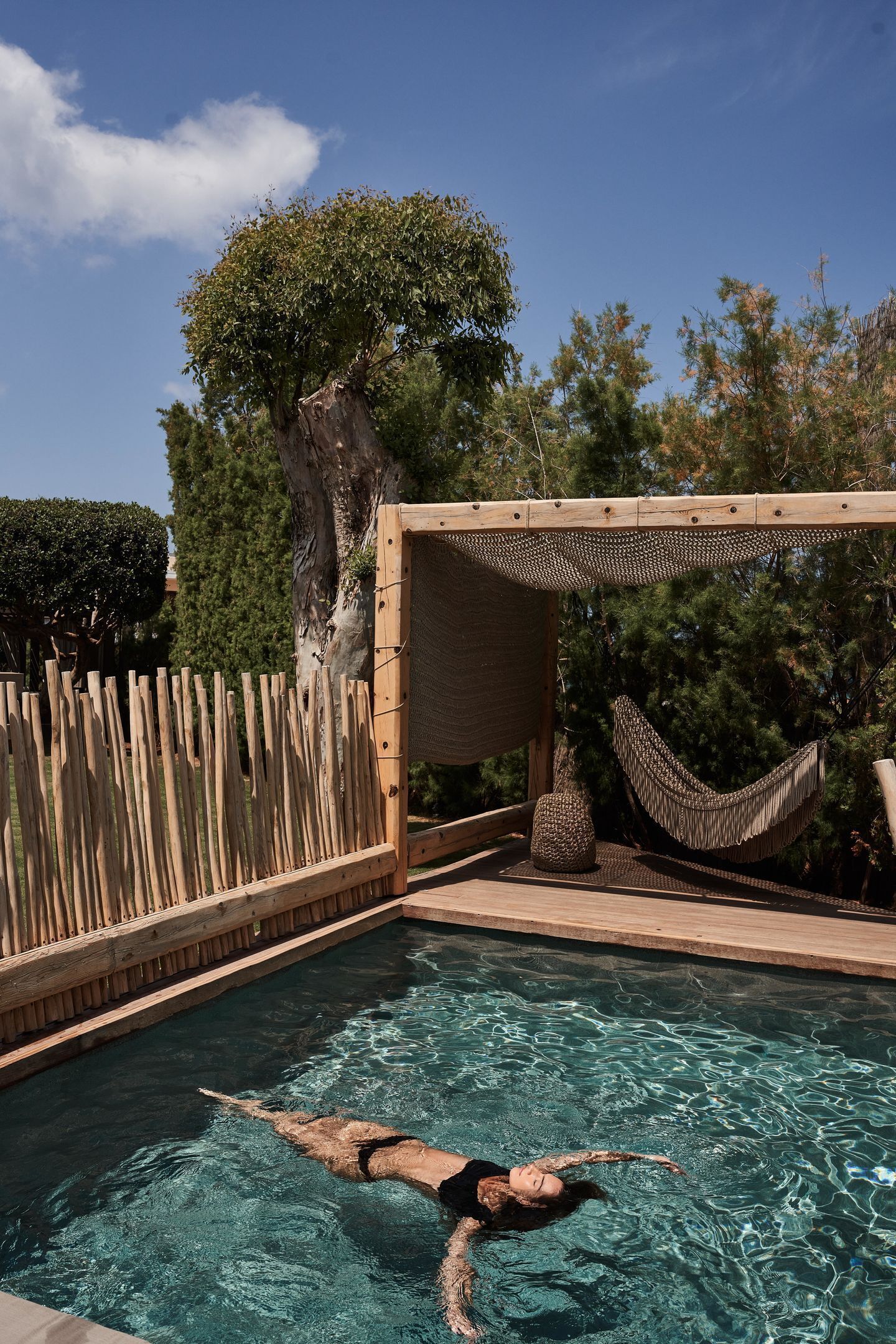 Un área de piscina con terraza de madera, una hamaca y un árbol grande.