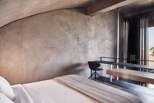 Una habitación moderna con una pared curva, una cama con ropa blanca, un tapiz tejido, una mesita pequeña y una silla.