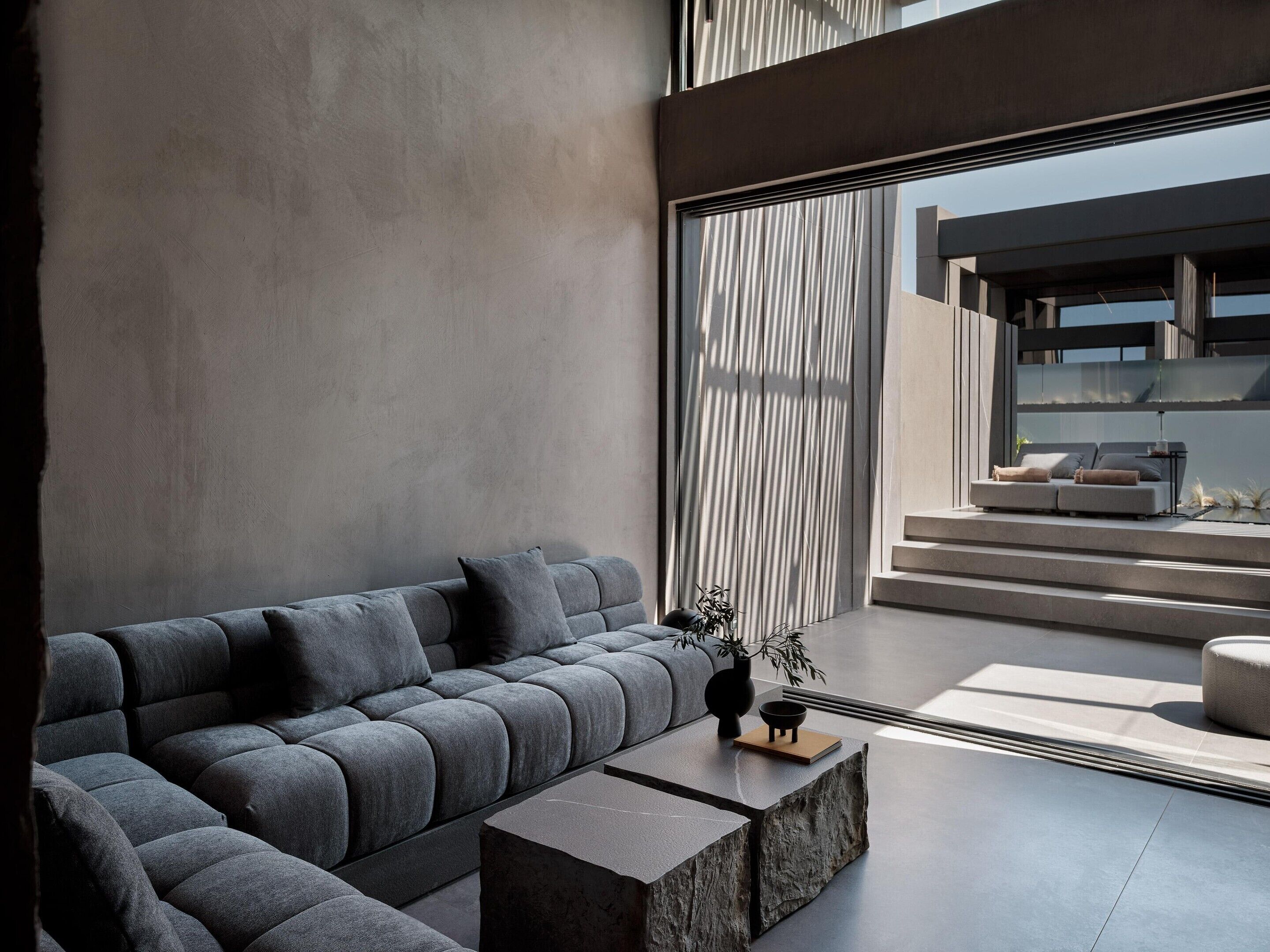 Un salon moderne avec un canapé d’angle, une table basse et une grande fenêtre dotée de stores verticaux.
