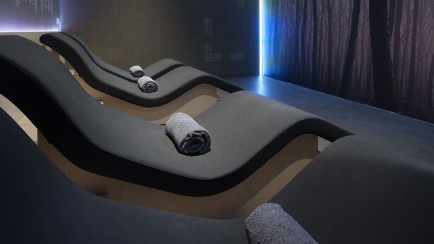 Ein moderner Wellnessraum mit drei Liegesesseln, jeweils mit einem Handtuch ausgestattet. Der Raum zeichnet sich durch ein minimalistisches Design mit dunkler Farbgebung und einem großen Bildschirm aus, der abstrakte Muster zeigt.