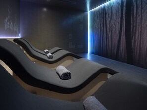A modern spa room with three reclining chairs, each with a towel. The room features a minimalist design with a dark color scheme and a large digital screen displaying abstract patterns. - NEMA Design Hotel & Spa - Small Luxury Hotels of the World (Hersonissos)