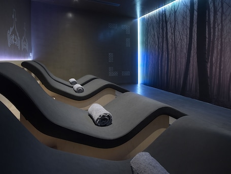A modern spa room with three reclining chairs, each with a towel. The room features a minimalist design with a dark color scheme and a large digital screen displaying abstract patterns.