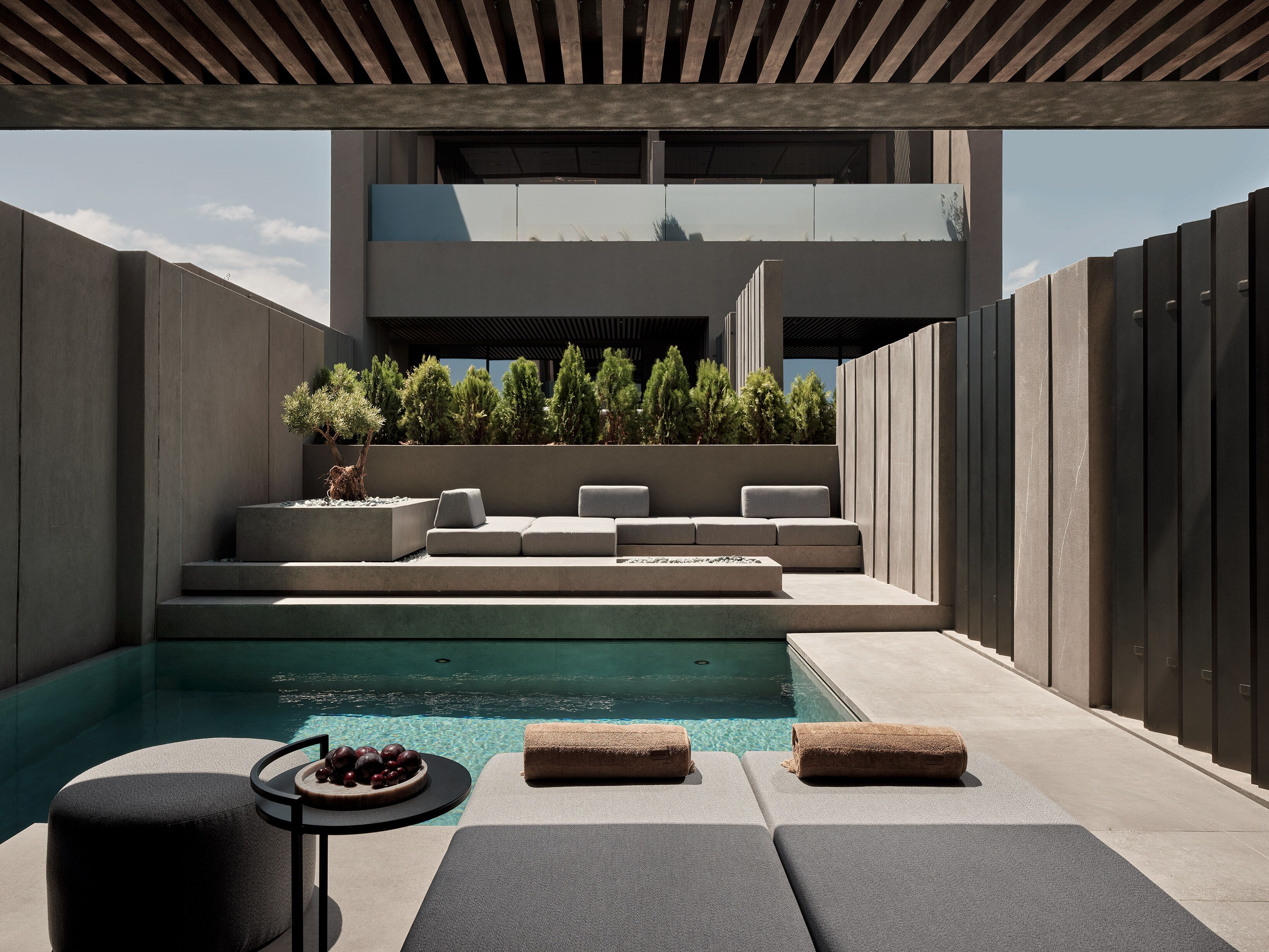 Un área de piscina moderna al aire libre con diseño minimalista, que incluye una piscina de hormigón, asientos y una pequeña mesa con frutas.