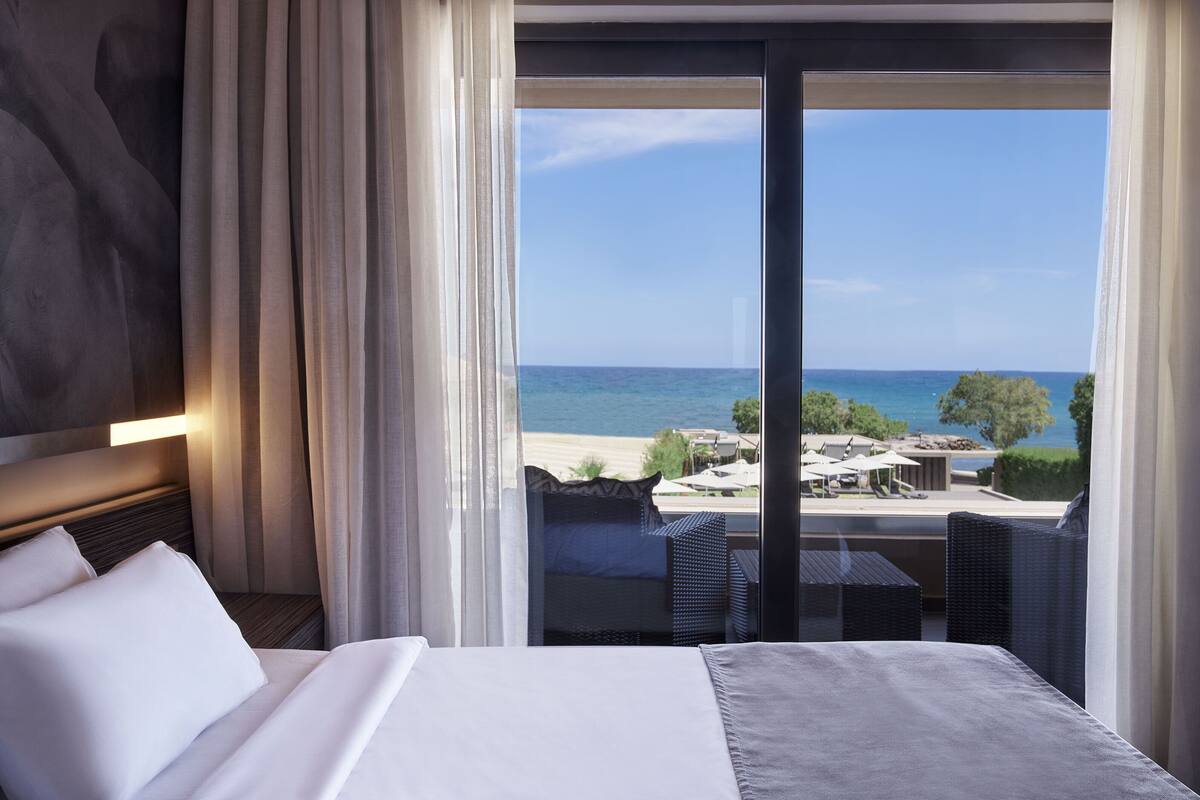 A hotel room with a large bed, a view of the beach, and a balcony with outdoor furniture.