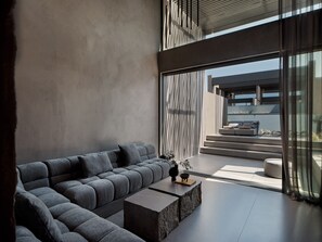 A modern living room with a sectional sofa, a coffee table, and a view through large windows to an outdoor seating area. - NEMA Design Hotel & Spa - Small Luxury Hotels of the World (Hersonissos)
