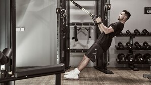 Un hombre realiza un ejercicio TRX en un gimnasio con jaulas de pesas y mancuernas.
