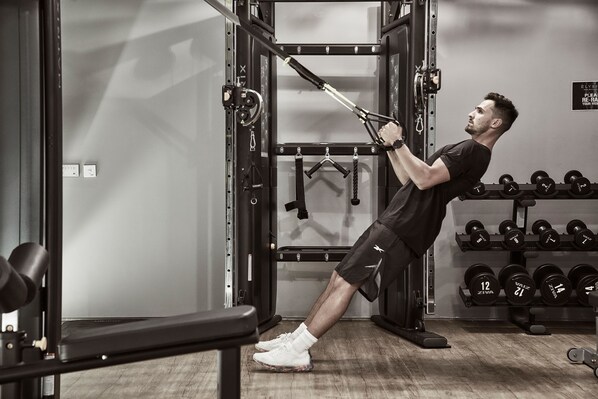 A man performing a TRX exercise in a gym with weight racks and dumbbells. - NEMA Design Hotel & Spa - Small Luxury Hotels of the World (Hersonissos)
