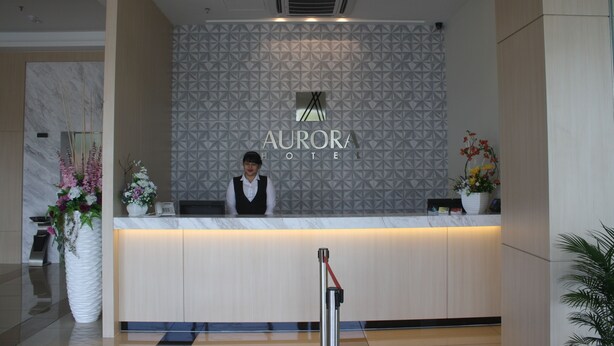 Una recepción de hotel con un papel tapiz de diseño geométrico, una recepcionista y un jarrón grande con flores.