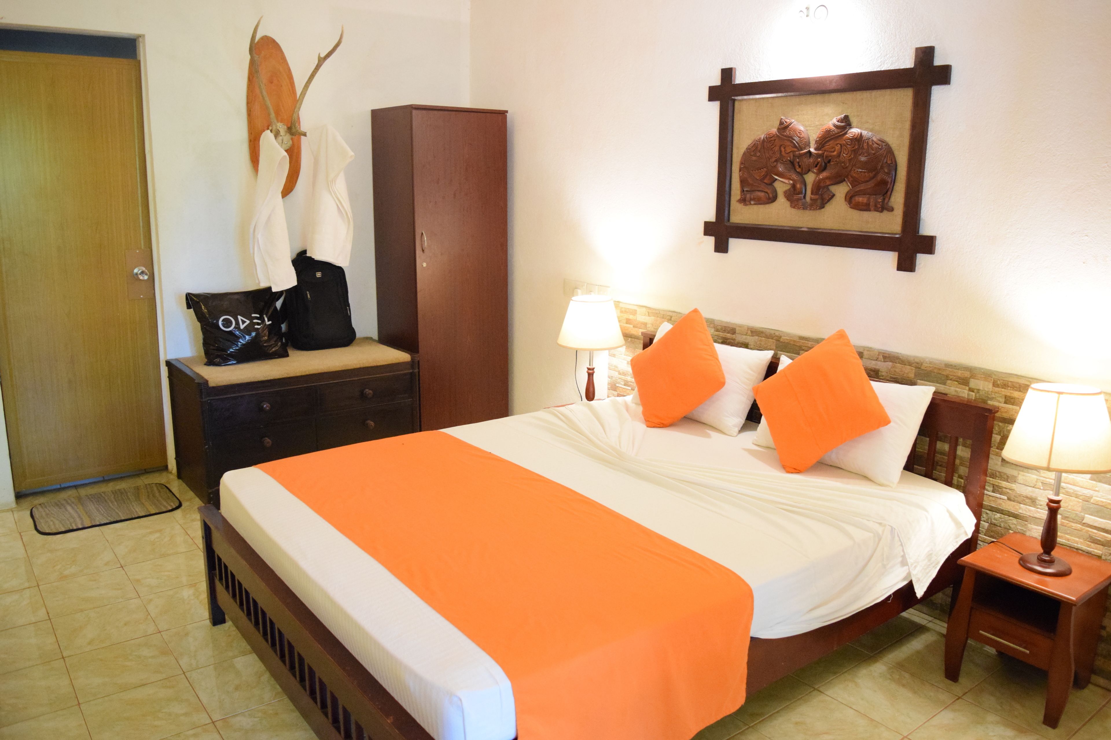 Habitación de hotel con cama, armario de madera, perchero con cabeza de venado, cuadro enmarco con dos gatos y una mesita con lámpara.