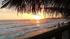A beach scene with palm leaves in the foreground, a sunset over the ocean, and a wooden fence along the shore. - Imagine Villa Hotel (Kamburugamuwa)