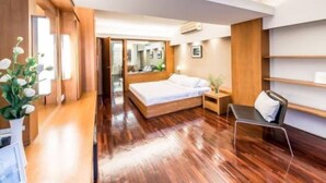 A modern hotel room with a wooden floor, a bed with white linens, a bedside table with a lamp, and a chair. - Laemtong Service Apartment (Si Racha)