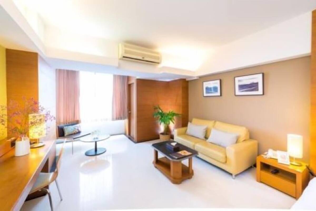 A modern hotel room with a sofa, a coffee table, a desk with a chair, and a bedside table with a lamp.