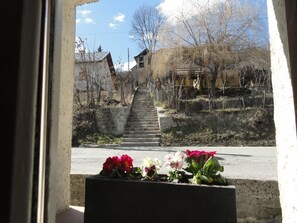 A view through a window of a stone staircase leading to a building with a tiled roof, surrounded by bare trees and a fence. - Hôtel Le Martagon (Villars-Colmars)