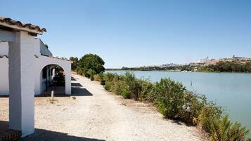 A gravel path leading to a white building with an archway, overlooking a calm river with a distant town on the opposite bank.