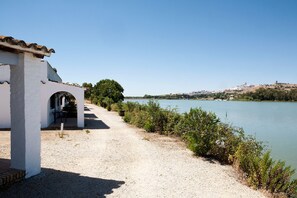 A gravel path leading to a white building with an archway, overlooking a calm river with a distant town on the opposite bank. - La Molinera Bungalows (Arcos de la Frontera)