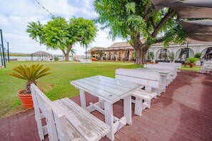 Un espace extérieur aménagé avec des tables et des bancs en bois peints en blanc, entouré de plantes en pot et d’arbres.